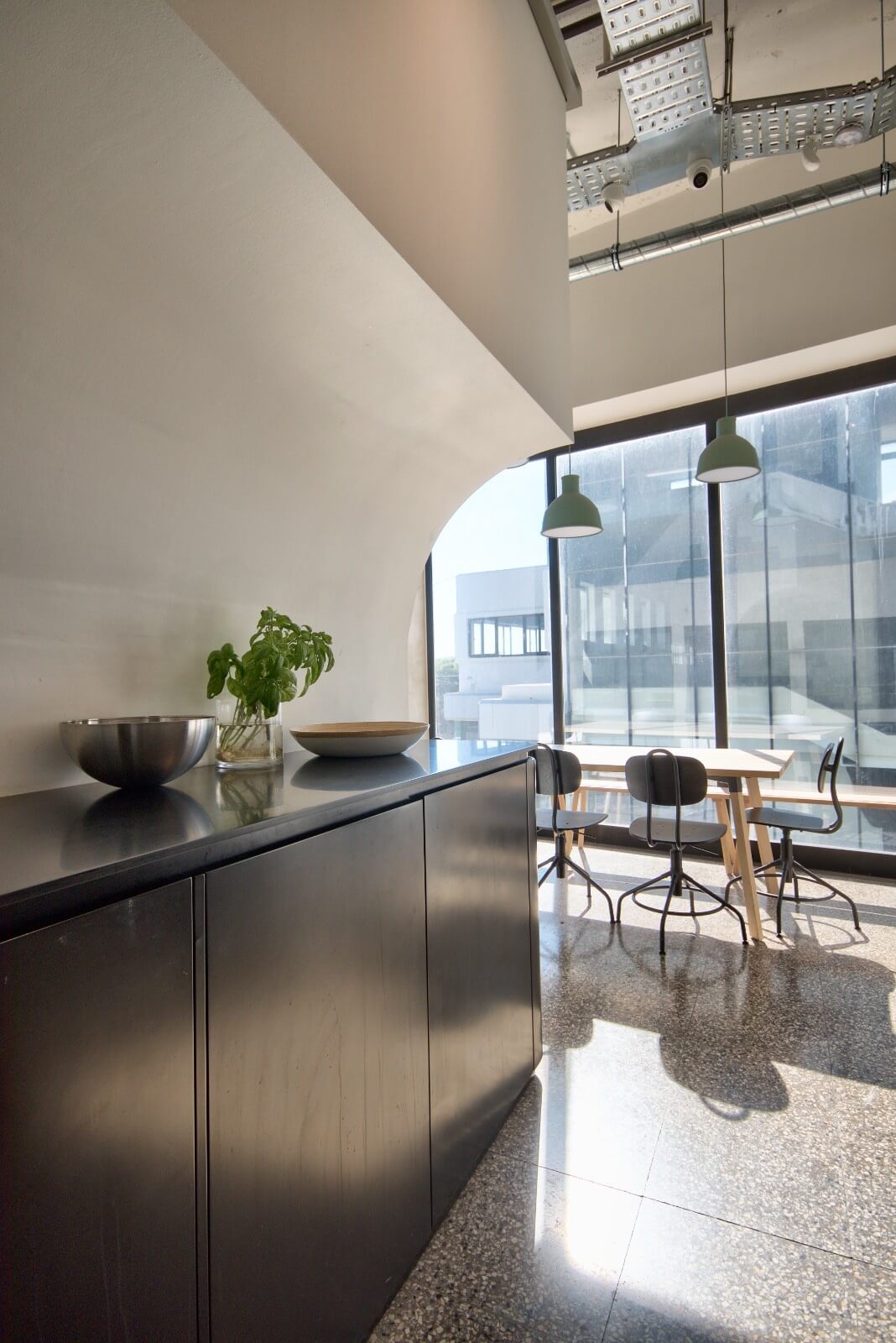 Bright office kitchenette and seating area with floor-to-ceiling windows, minimalist pendant lights, and a sleek black cabinet topped with bowls and a basil plant, set against Steel Grey tiled flooring.