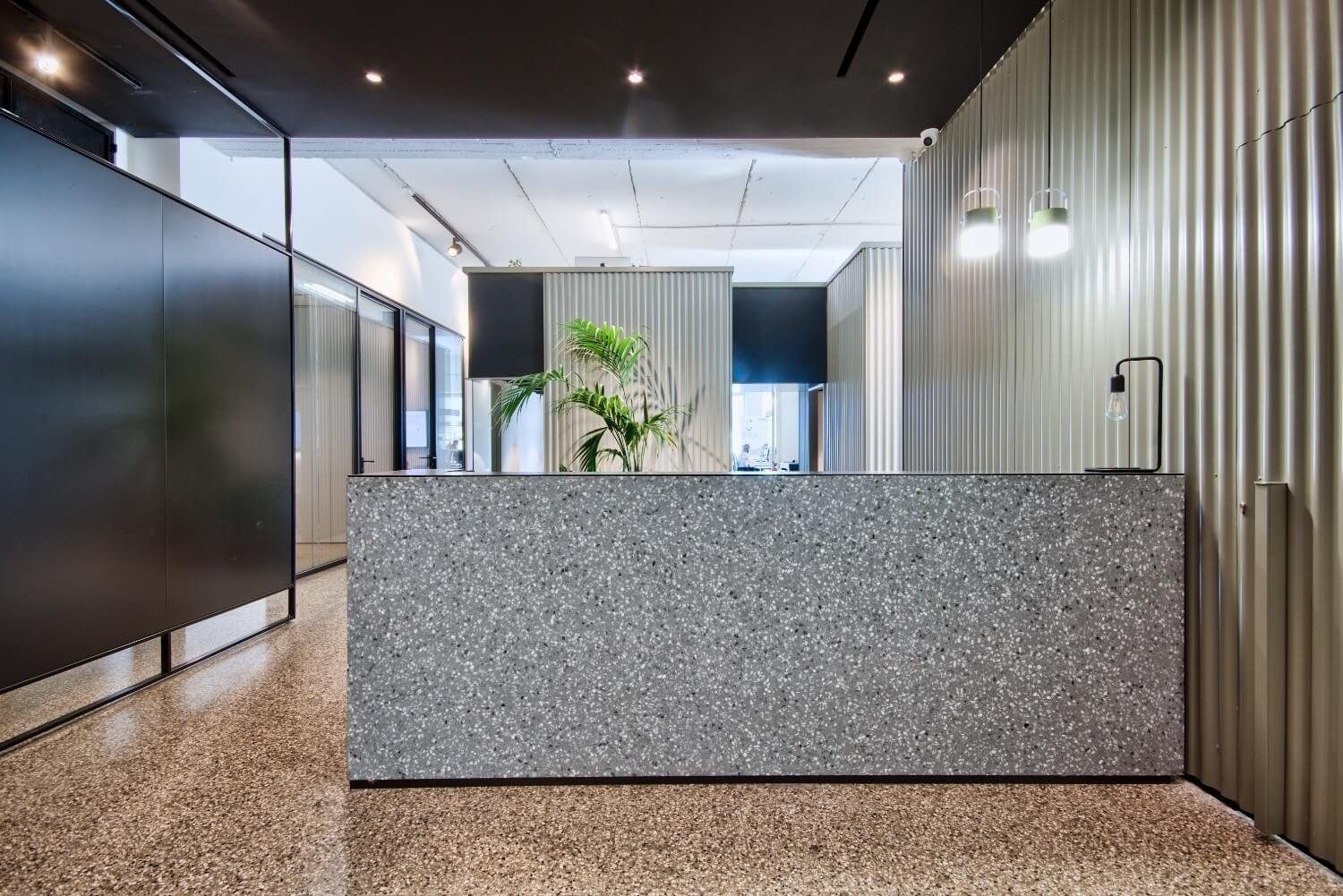 Front view of a contemporary office reception desk clad in Steel Grey terrazzo, with corrugated panel walls, hanging lights, and in-situ polished flooring from the Minima Collection.