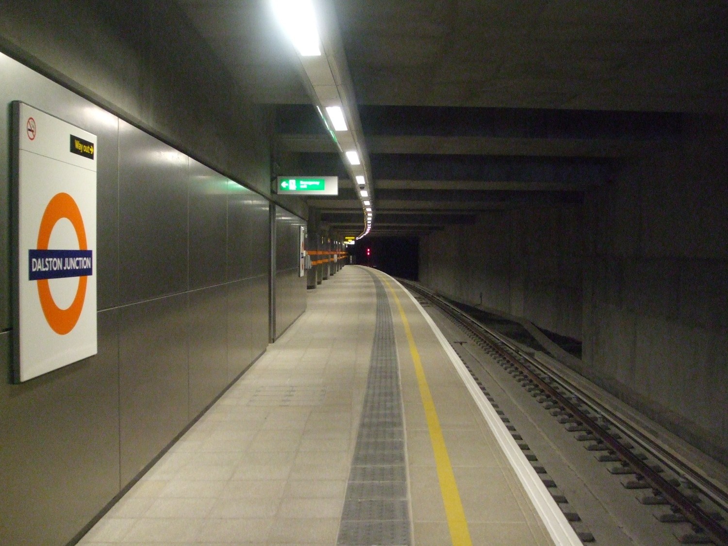 Wide underground platform view at Dalston Junction station with polished grey terrazzo flooring, a curved track, and overhead lighting leading into a tunnel.