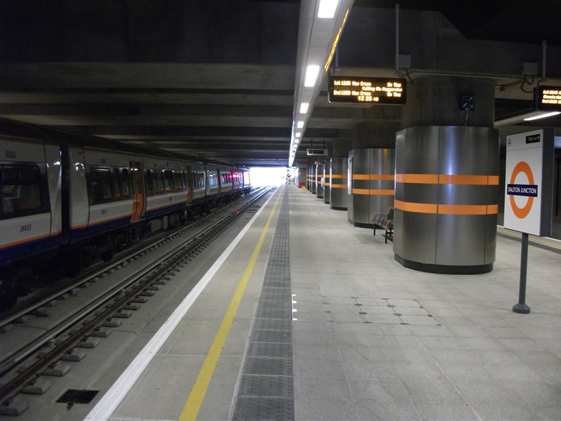 Spacious underground platform at Dalston Junction station with cylindrical columns clad in brushed metal and orange stripes, digital departure boards, and a London Overground train on the left.