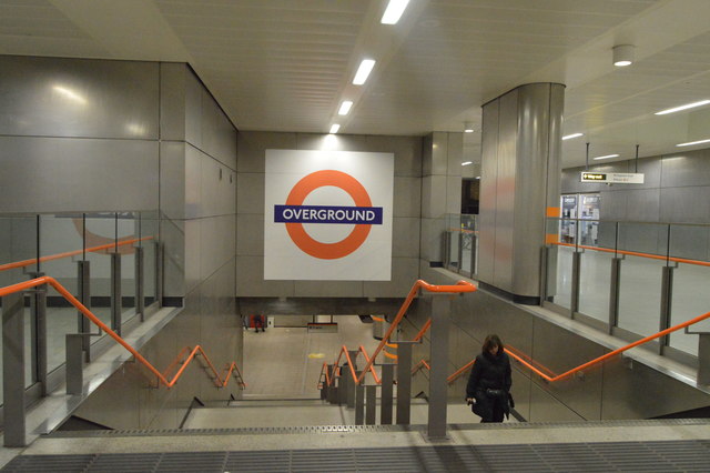 Entrance view at a London Overground station featuring a prominent Overground roundel sign on a tiled wall, flanked by grey interiors and orange handrails on descending stairs.