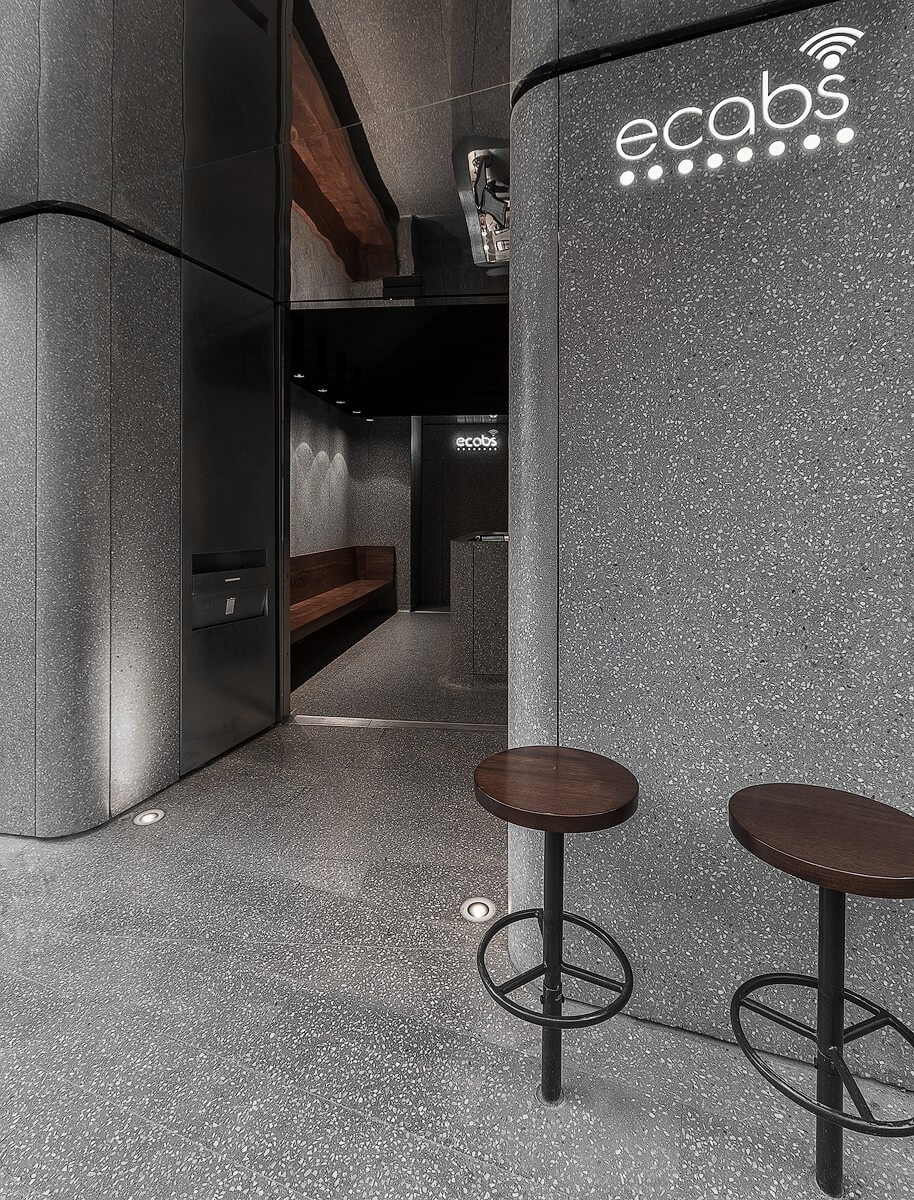 Side view of the Ecabs entrance showcasing illuminated branding on terrazzo-clad curved walls, two bar stools, and a glimpse of the walnut bench inside.