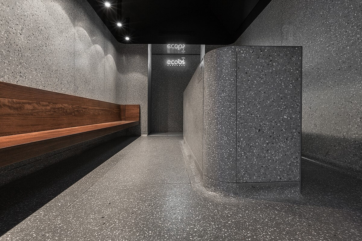 Low-angle view of the Ecabs interior showing the walnut seating area alongside a terrazzo-clad curved reception desk under minimal black ceiling lighting.