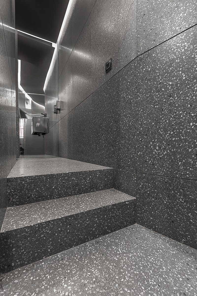 Minimalist corridor with terrazzo steps and walls at Ecabs, leading to a well-lit area with suspended ceiling panels and restroom signage.