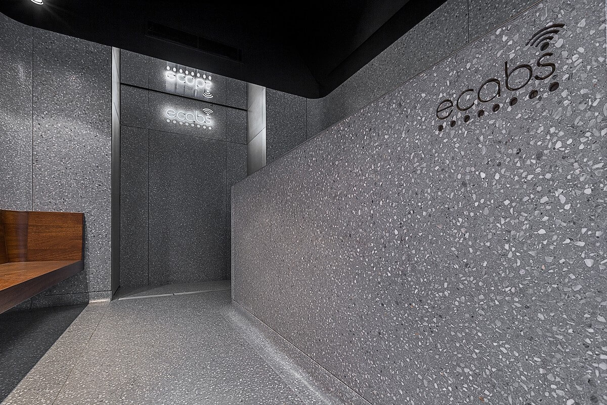Interior view of the Ecabs reception space with a curved terrazzo reception desk and backlit wall signage, adjacent to a walnut bench and dark ceiling panels.