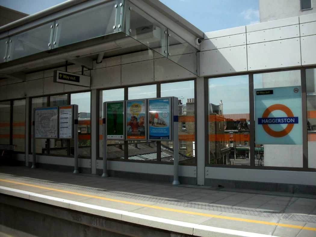 Haggerston station platform with Overground signage, poster displays, and full-height glass windows revealing East London rooftops in the background.