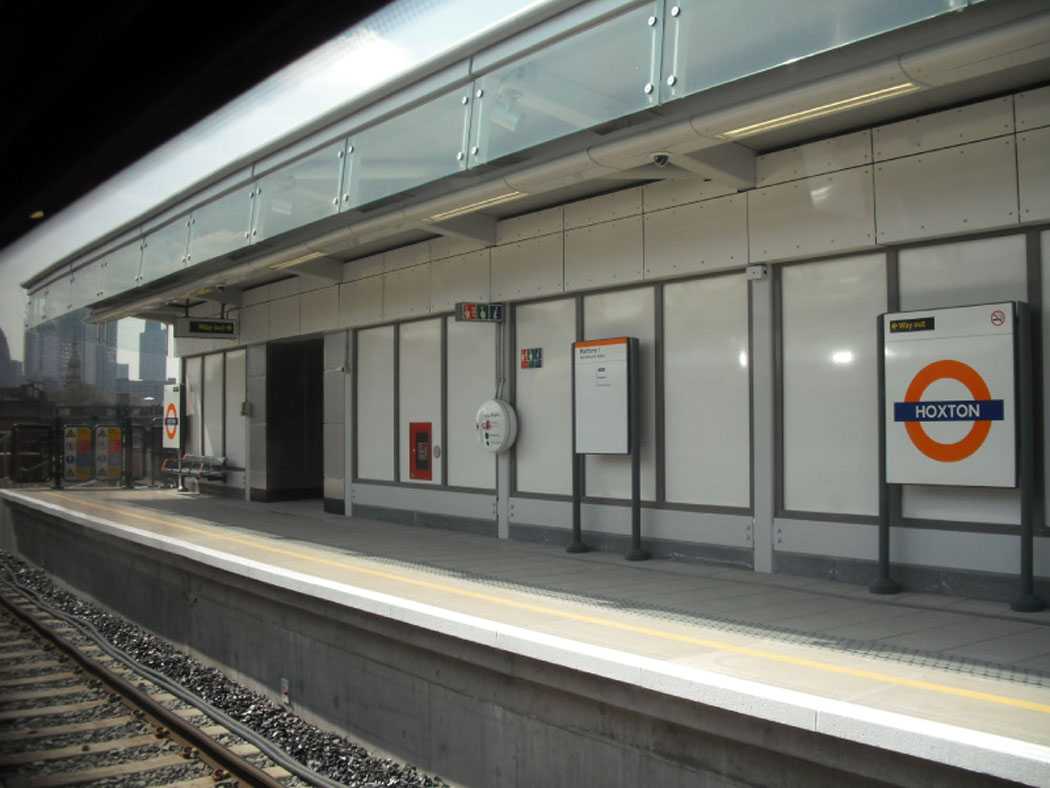View from inside a train at Hoxton station platform, showing window-lined walls, benches with orange armrests, and clear visibility of the Overground roundel.