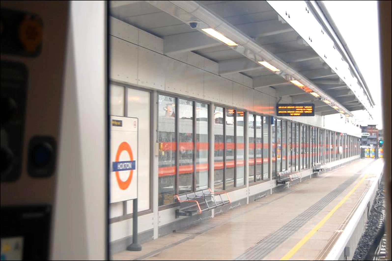 Interior ticket barrier area at Hoxton Overground station with accessible gates, sleek grey walls, a blue service counter, and escalators in the background.