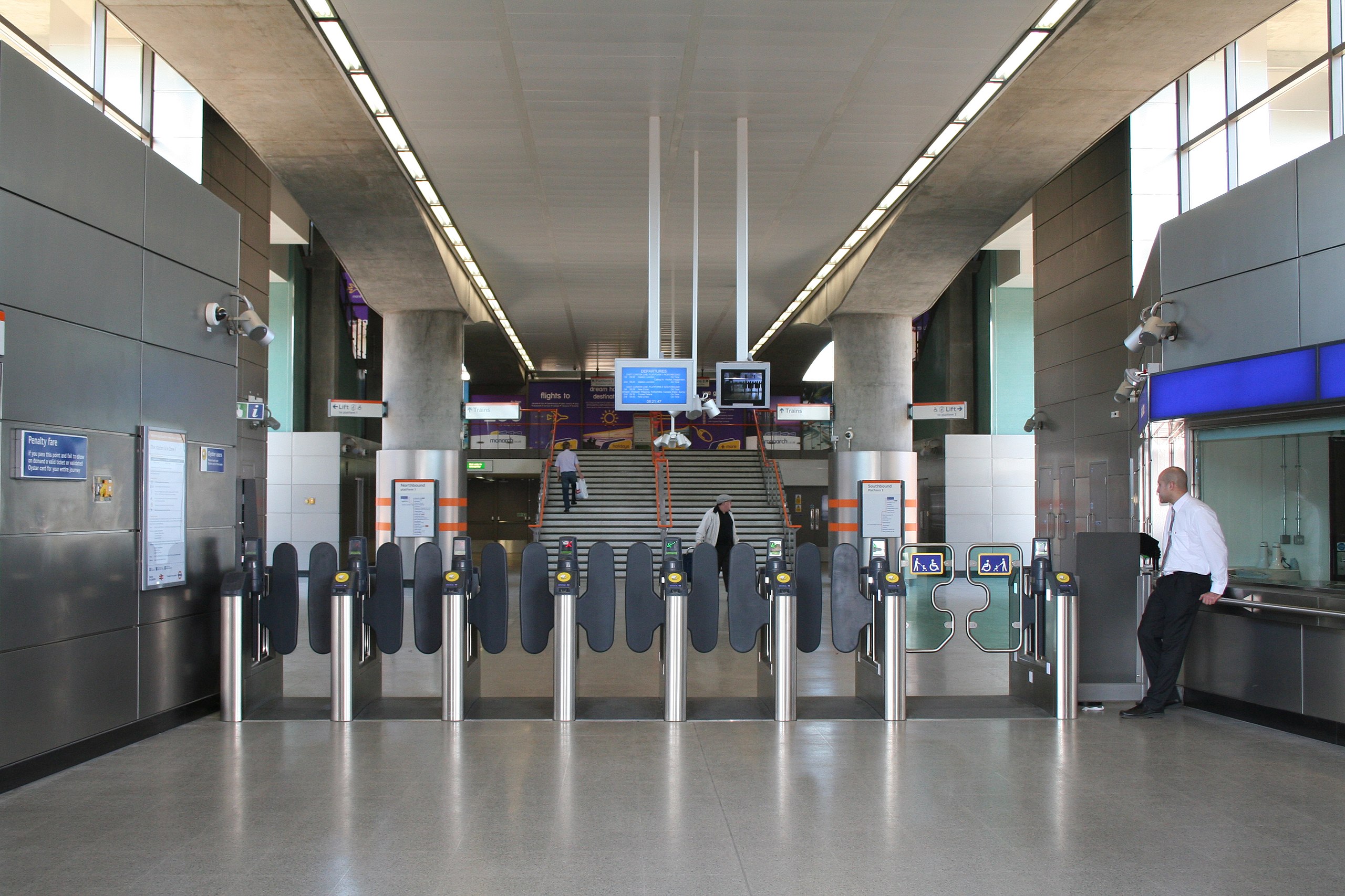 Shoreditch High Street Overground station platform with metallic benches, a visible roundel sign, and a yellow-and-blue London Overground train moving at high speed.