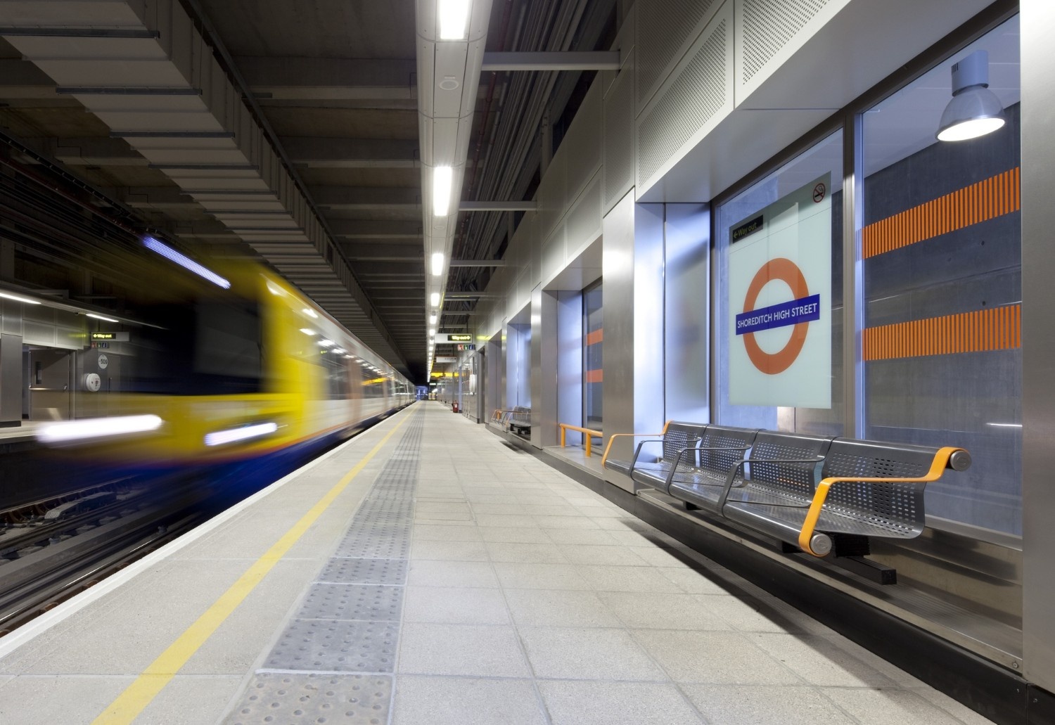 Modern and well-lit platform at Shoreditch High Street station with perforated benches, terrazzo-like flooring, and the station’s roundel sign on a large wall panel.