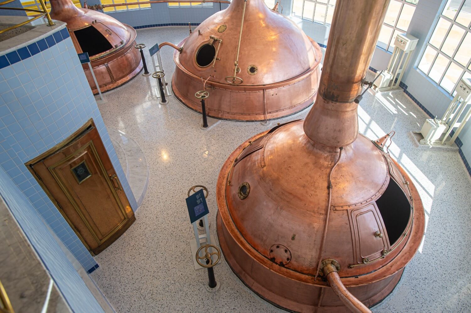 Overhead view of a brewery exhibit featuring large, restored copper brewing kettles placed on polished terrazzo flooring with speckled blue chippings.