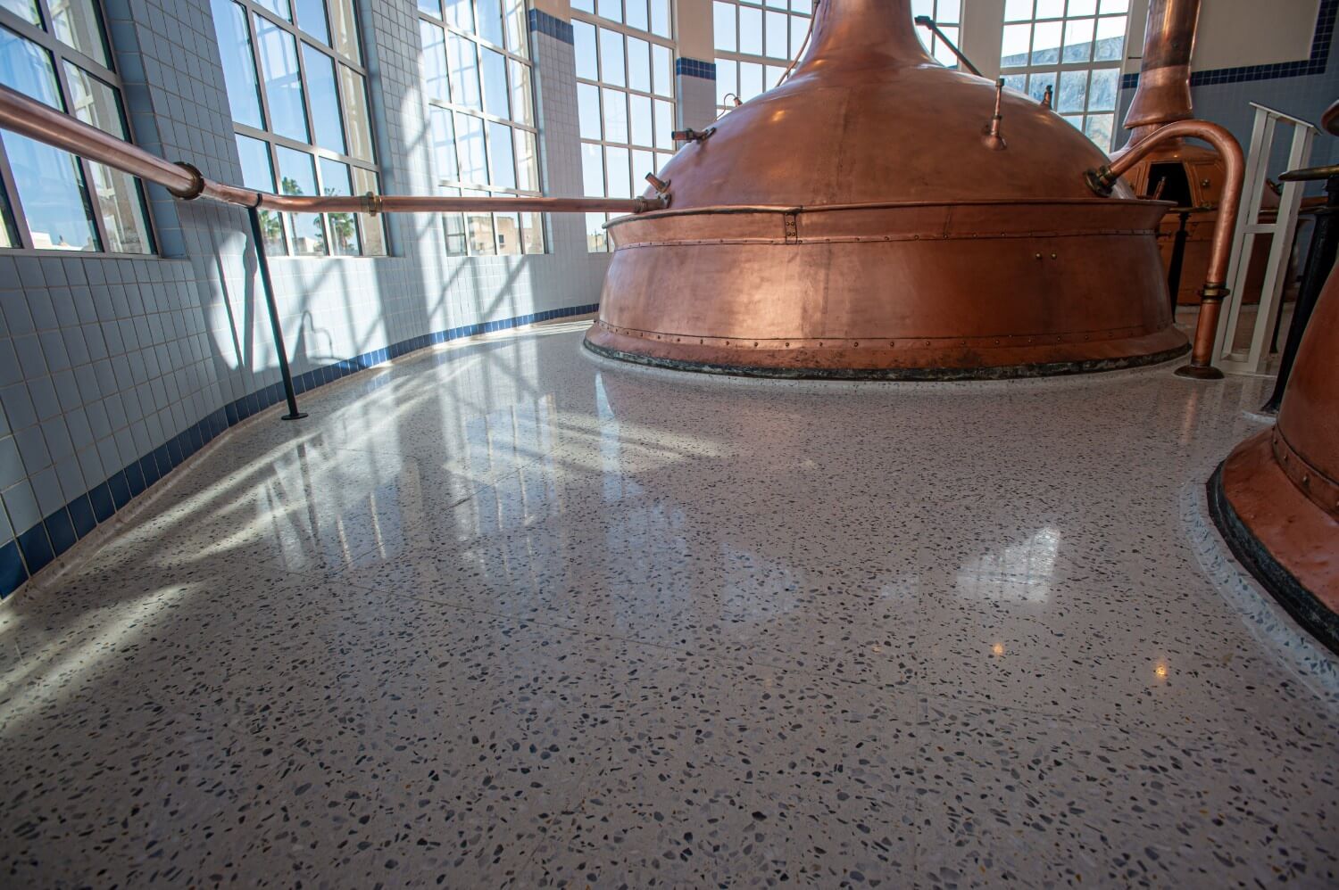 Side view of a copper brewing vat positioned near tall curved windows, with natural light reflecting on the newly installed terrazzo floor.