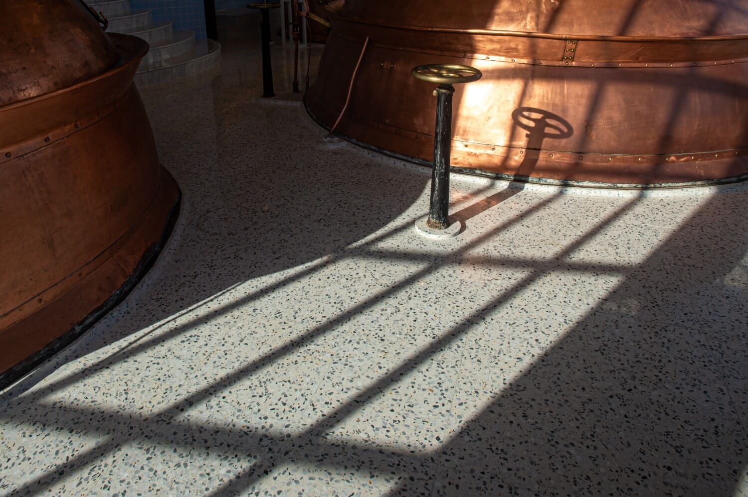 Sunlight casts shadows across the terrazzo floor in the Farsons Brewhouse, illuminating the large copper brewing kettles. The polished finish and aggregate mix in the flooring are clearly visible.