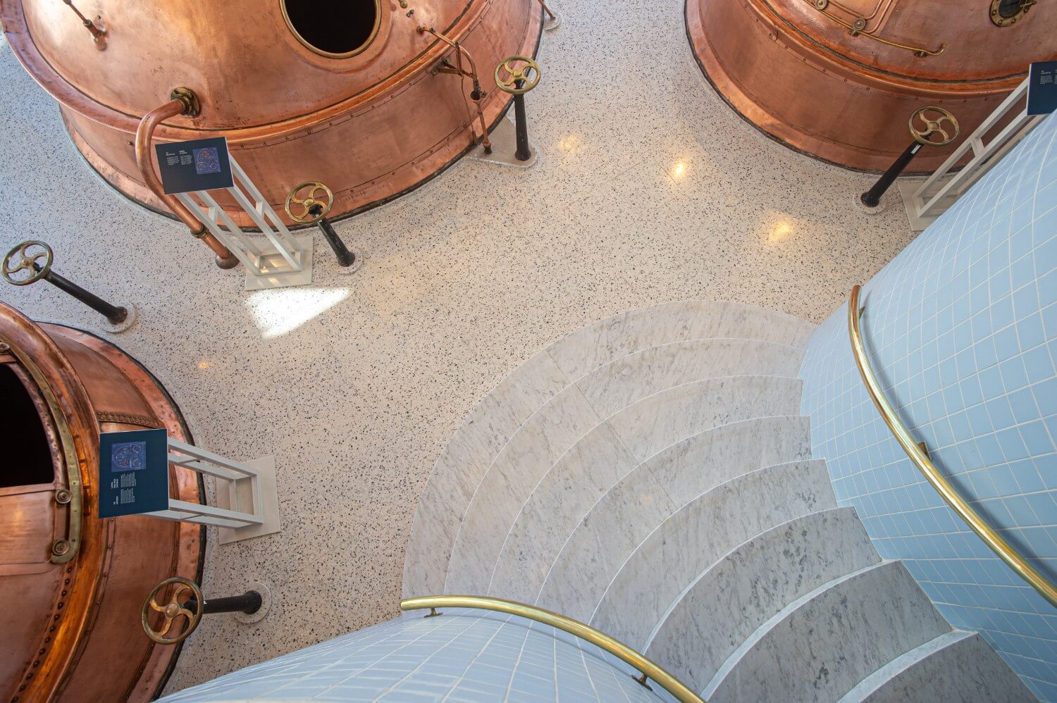 Top-down view of the exhibit area with circular stairs leading to the terrazzo floor, surrounded by copper brewing vats and display signage.
