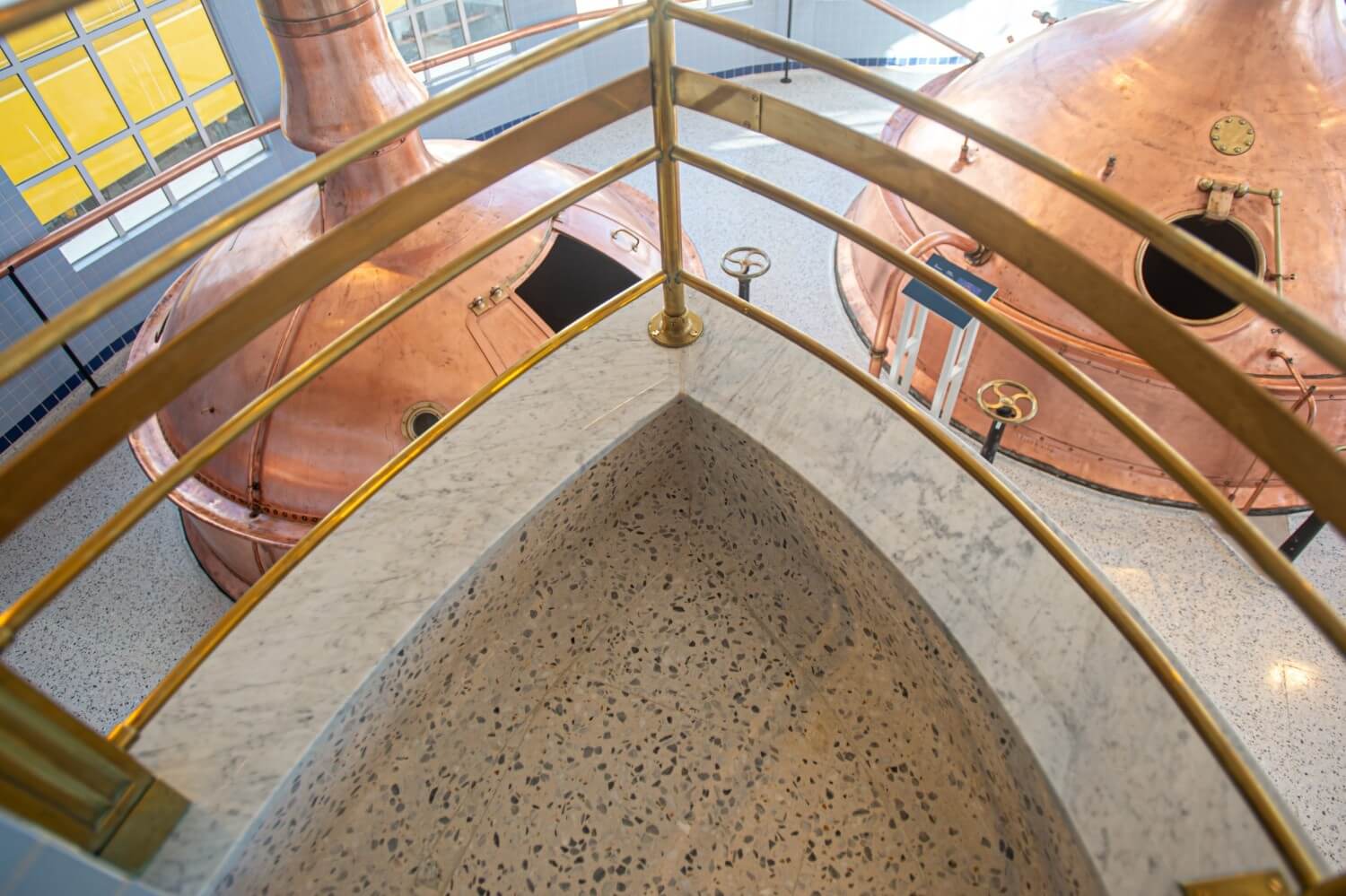 Top view of a triangular landing at the edge of the Farsons Old Brewhouse mezzanine, finished in terrazzo with a marble border and brass handrail, overlooking copper vats below.