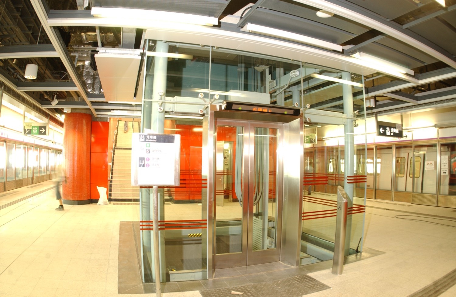 Glass elevator enclosure at Kowloon Station platform level, framed by red tiled walls and steel columns, with train doors visible in the background.