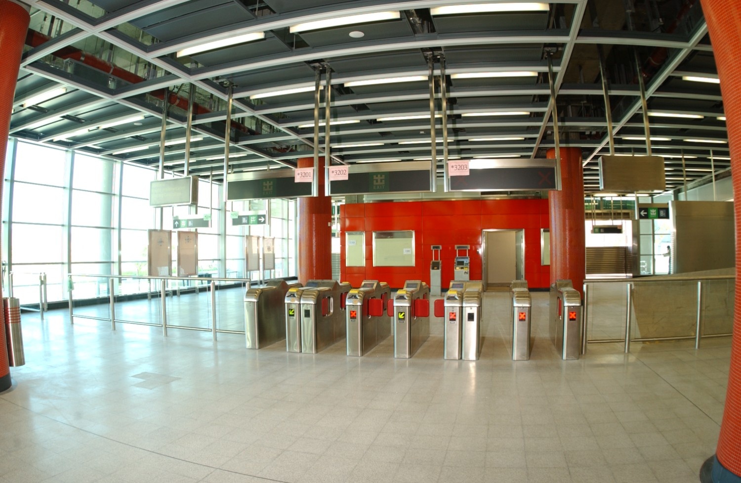 Interior view of Kowloon Station’s ticket barrier area featuring modern red wall panels, stainless steel turnstiles, and a glass façade allowing natural light to flood the space.